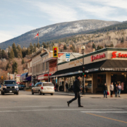 A person walks across the Baker Street in downtown Nelson, BC. There is a slight skiff of snow on the mountain behind buildings.